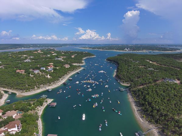 Aerial drone view of a summer lake cove with dozens of boats clustered on turquoise water, surrounded by tree‑covered shores and waterfront homes beneath a bright blue sky