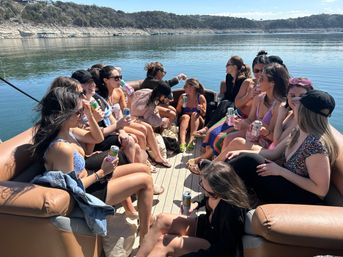 Group of friends enjoying a sunny pontoon boat outing on a calm lake, sitting on cushioned seats in swimsuits and summer clothes while sipping canned drinks, with a rocky, tree-lined shoreline in the background.