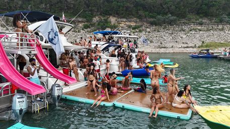 Busy summer lake party with anchored boats, pink water slides and a crowded floating dock of people in swimsuits by a rocky tree-lined shoreline.