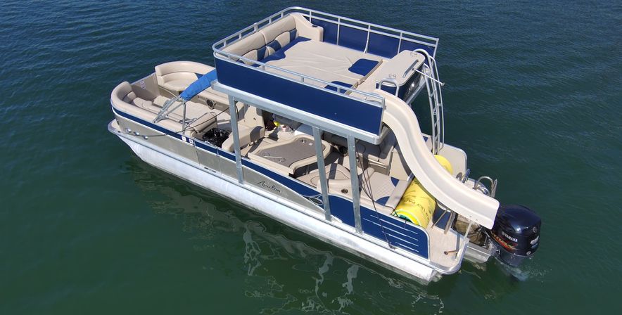 Aerial view of a blue-and-white pontoon boat with an upper deck and curved water slide floating on a calm green lake, ready for summer fun