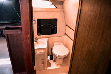 Compact boat cabin bathroom (marine head) with white toilet, small sink, mirrored hatch and warm wood paneling — cozy, space‑saving yacht head.
