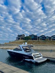 Sleek black-and-white motorboat moored at a lakeside dock with modern waterfront homes on a rocky bluff beneath a dramatic rippled cloud sky.