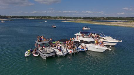 Summer boat party on a lake: yachts, pontoons and jet skis rafted together with crowds of people, blue water, sandy shoreline and sunny sky.