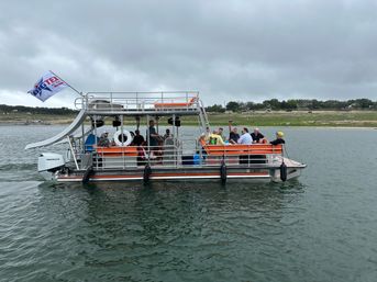 Pontoon party boat with orange benches and a waterslide carrying a group on a calm recreational lake under a cloudy sky, grassy shoreline in the background.