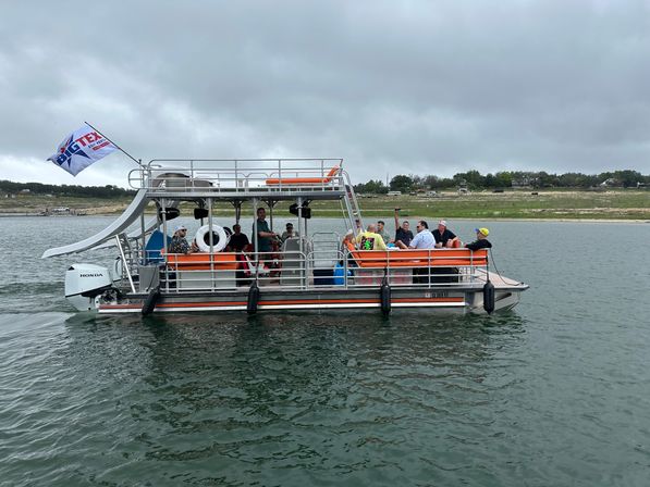 Pontoon party boat with orange benches and a waterslide carrying a group on a calm recreational lake under a cloudy sky, grassy shoreline in the background.