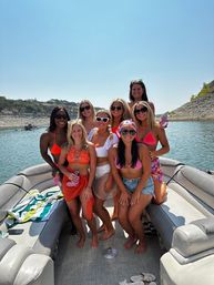 Eight friends posing on a pontoon boat at a sunny lake, wearing colorful swimsuits and sunglasses with a rocky shoreline and clear blue sky in the background