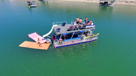 Aerial view of a double-deck pontoon boat partying on a green lake with people sunbathing, a white slide and pink and orange floating mats attached for swimming and lounging near the shoreline.