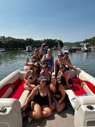 Group of friends in swimsuits smiling and holding drinks on a red-and-white pontoon boat at a sunny summer lake party with boats and tree-lined shore in the background.