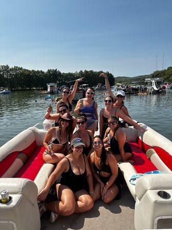 Group of friends in swimsuits smiling and holding drinks on a red-and-white pontoon boat at a sunny summer lake party with boats and tree-lined shore in the background.