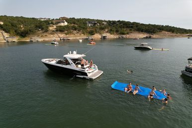 Summer boating scene on a calm lake: a black-and-white motorboat with friends aboard anchored near a wooded shoreline while people relax and swim on a large blue floating mat, other boats and lakeside homes in the background.