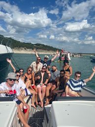 Group of friends on a motorboat enjoying a sunny summer lake party with swimsuits and red cups, anchored near a rocky shoreline and other party boats under a blue sky with puffy clouds.