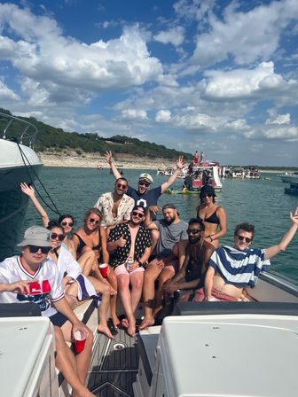 Group of friends on a motorboat enjoying a sunny summer lake party with swimsuits and red cups, anchored near a rocky shoreline and other party boats under a blue sky with puffy clouds.