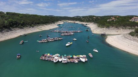 Aerial view of a summer lake cove with clusters of party boats and floating rafts anchored in turquoise water, wooded shoreline and hillside homes under a partly cloudy sky.