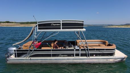 Black-and-silver pontoon boat with tan bench seating and a waterslide, a person at the helm under a double-decker canopy floating on a sunny blue lake with a distant sandy shoreline.