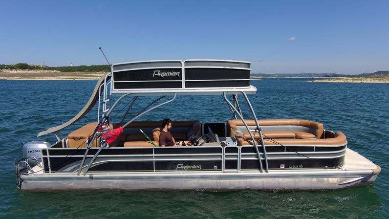 Black-and-silver pontoon boat with tan bench seating and a waterslide, a person at the helm under a double-decker canopy floating on a sunny blue lake with a distant sandy shoreline.