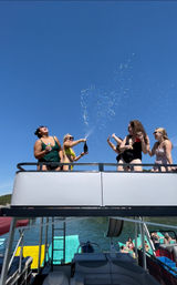 Summer lake party: friends laughing as champagne sprays from the upper deck of a pontoon boat under a clear blue sky.