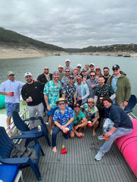 Large group of men on a pontoon boat at a lake cove, many wearing Hawaiian shirts and caps and holding red cups, surrounded by rocky shoreline, hills and a cloudy sky