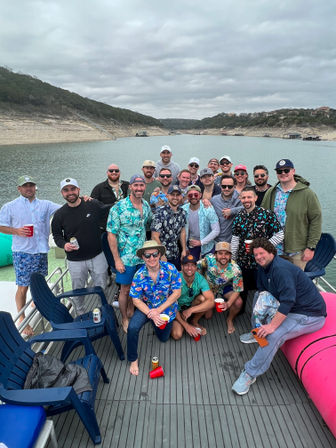 Large group of men on a pontoon boat at a lake cove, many wearing Hawaiian shirts and caps and holding red cups, surrounded by rocky shoreline, hills and a cloudy sky
