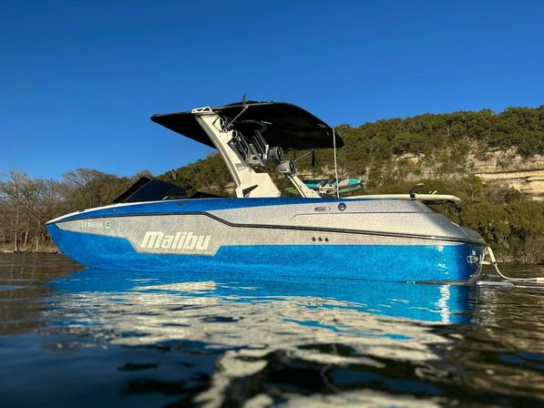 Sparkling blue-and-silver wakeboard boat with black tower and canopy floating on a calm lake near a wooded shoreline and rocky bluff under a clear blue sky.