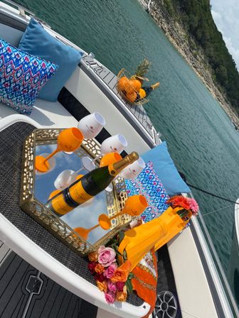 Boat deck picnic on a turquoise lake near a rocky shore — mirrored tray with a champagne bottle and bright orange goblets, a fruit basket with pineapple and oranges, colorful patterned pillows and fresh flowers.