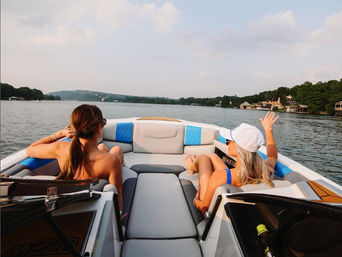 Two friends lounging at the bow of a speedboat on a calm lake, waving toward tree-lined shoreline and waterfront homes under a hazy summer sky.