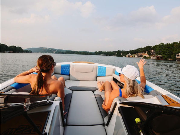 Two friends lounging at the bow of a speedboat on a calm lake, waving toward tree-lined shoreline and waterfront homes under a hazy summer sky.