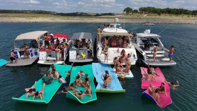 Summer lake boat party with multiple motorboats tied together near a rocky shoreline, people socializing on decks and lounging on colorful foam float mats on clear blue water.