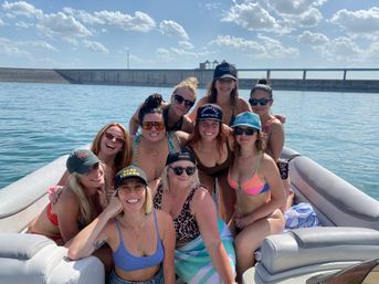 Smiling group of friends in bikinis and caps lounging on a pontoon boat on a sunny lake, with a concrete dam and fluffy clouds on the horizon.