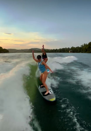 Two riders wakesurfing on a lake at sunset, one in a blue life vest carving the boat wake with a waving companion behind, tree-lined shore and distant bridge