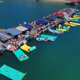 Aerial view of a lively lake dock party with a row of pontoon boats tied together, colorful floating mats and slides on teal water, people relaxing and socializing on a sunny summer day.