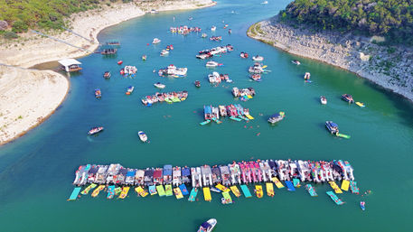 Aerial view of a summer lake cove crowded with dozens of pontoon and pleasure boats lined in a long row, colorful floating mats and inflatables on turquoise water between rocky, tree-covered shorelines