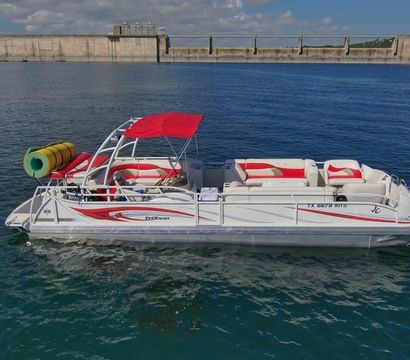 White-and-red pontoon boat with a red canopy and rolled-up inflatables floating on clear blue reservoir water in front of a long concrete dam under a partly cloudy sky