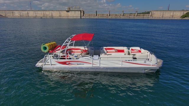 White-and-red pontoon boat with a red canopy and rolled-up inflatables floating on clear blue reservoir water in front of a long concrete dam under a partly cloudy sky