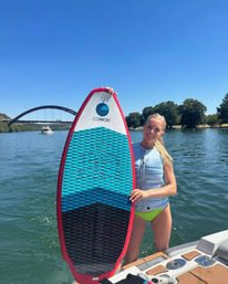 Smiling woman in a life vest on a boat holding a blue-and-black wakesurf board with a red edge on a sunny lake, with an arched bridge and tree-lined shore in the background.
