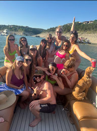 Group of friends in colorful swimsuits smiling and posing on a crowded pontoon boat with a golden dog and drinks, clear blue sky and rocky lake shoreline with houses in the background.
