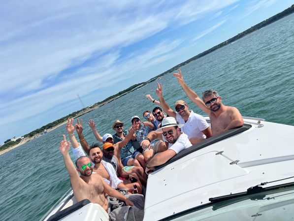 Group of friends cheering on a white motorboat on a sunny summer day in coastal waters with a sandy shoreline in the background.