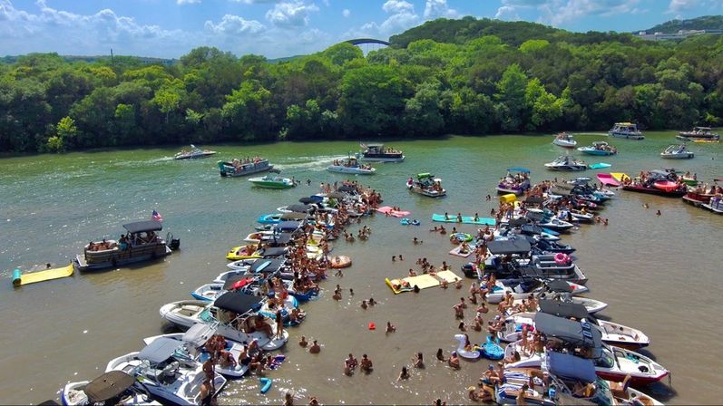 Aerial view of a crowded summer river float: dozens of boats and pontoons clustered in shallow water with people swimming, lounging on floating mats and tubes near a green tree-lined shore under a sunny blue sky.