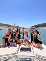 Group of friends in swimsuits laughing on the bow of a boat during a sunny summer lake day with wooded shores and other boats in the background.