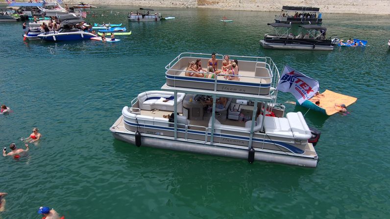 Double-decker pontoon boat on a green lake with people socializing on the upper deck, swimmers and nearby boats and floaters, rocky shoreline in the distance — sunny summer boating party.