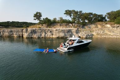 Motorboat anchored near limestone cliffs on a calm lake, people relaxing on the swim platform and lounging on a large blue floating mat, summer shoreline with trees.