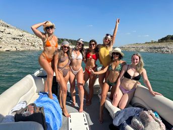 Eight friends in colorful bikinis and summer hats smiling and posing on a pontoon boat at a rocky lake shoreline under a clear blue sky