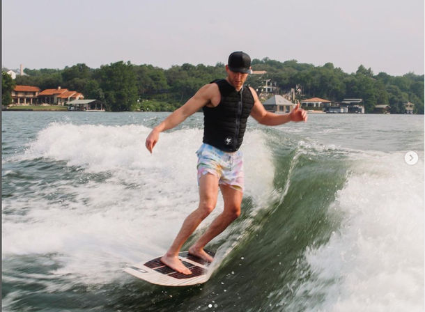 Wakesurfer on a lake cutting a green wake near a tree-lined shoreline with lakeside homes, wearing a black life vest, cap, and pastel shorts