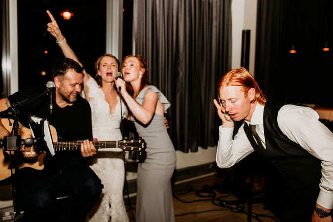 Indoor evening wedding reception sing-along with live acoustic guitar: woman in white lace dress and woman in grey gown share a microphone as a seated guitarist plays and a suited guest leans in, dancing and smiling.
