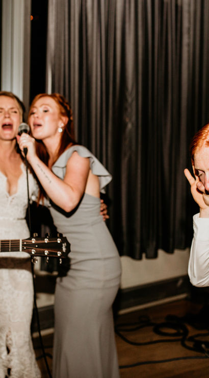 Indoor evening wedding reception sing-along with live acoustic guitar: woman in white lace dress and woman in grey gown share a microphone as a seated guitarist plays and a suited guest leans in, dancing and smiling.