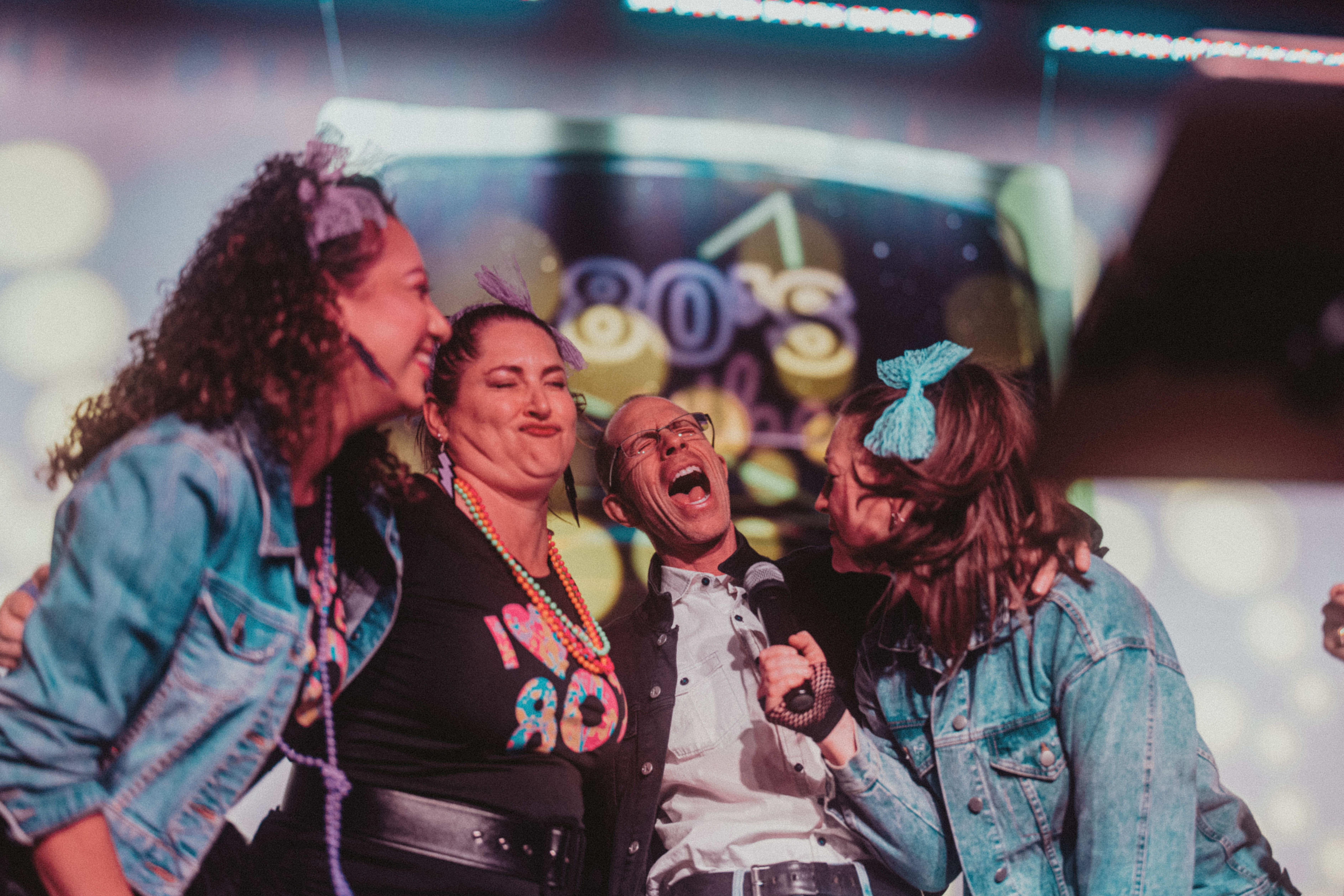 Four friends in 1980s-inspired outfits laughing and singing into a microphone on stage with a neon '80s' backdrop and colorful retro party lighting.