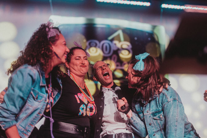 Four friends in 1980s-inspired outfits laughing and singing into a microphone on stage with a neon '80s' backdrop and colorful retro party lighting.