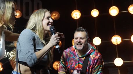 Female singer holding a microphone smiling at a male guitarist in a colorful hoodie on a live music stage with warm round stage lights