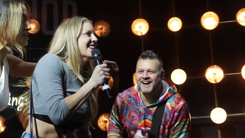 Female singer holding a microphone smiling at a male guitarist in a colorful hoodie on a live music stage with warm round stage lights