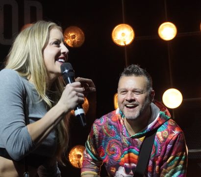 Female singer holding a microphone smiling at a male guitarist in a colorful hoodie on a live music stage with warm round stage lights