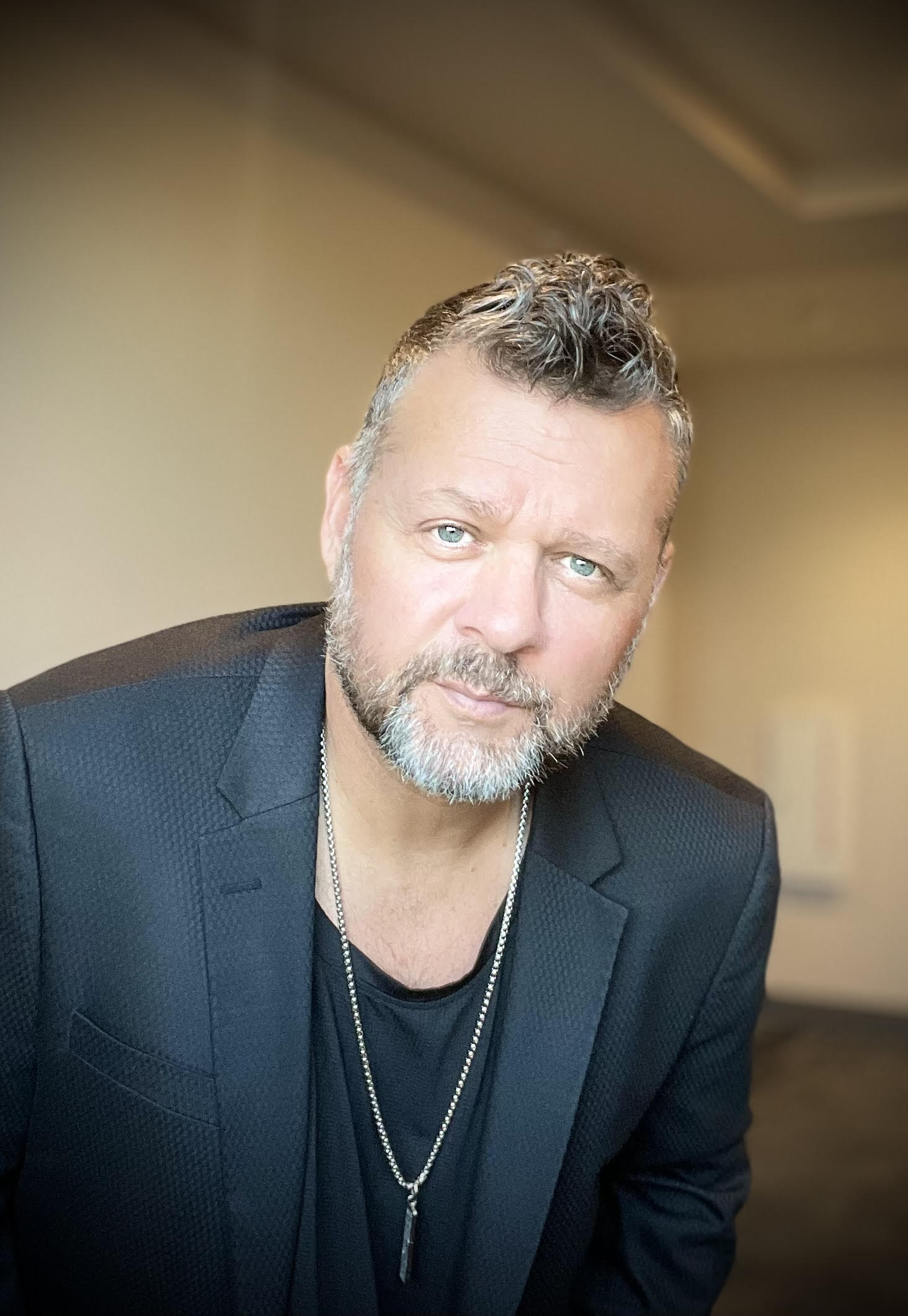 Confident indoor headshot of a middle-aged man with salt-and-pepper beard and short curly hair, wearing a black blazer and pendant necklace against a soft neutral background.